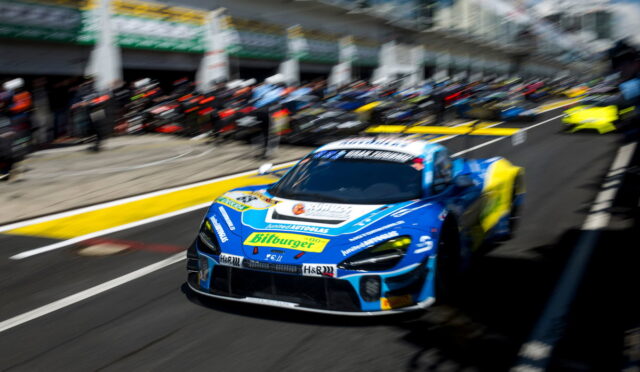 Blue race car with sponsor logos speeding from the pit lane amid a blurred crowd and garages in the background.