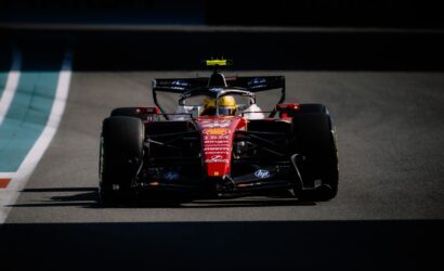 Red Ferrari Formula 1 car on a race track, driver wearing a yellow helmet, sponsor logos visible on the chassis From front view.