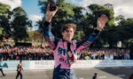 Racing driver in a pink suit raises arms in triumph while taking a selfie with the cheering crowd behind him.