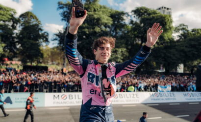 Racing driver in a pink suit raises arms in triumph while taking a selfie with the cheering crowd behind him.