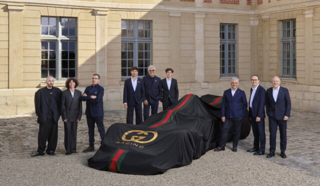 Group of formally dressed people standing around a covered race car in a courtyard, awaiting reveal.
