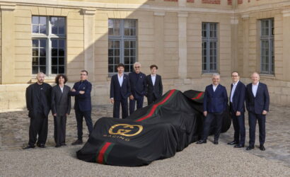 Group of executives in suits posing beside a race car under a black cover with red and green stripes, in a stone courtyard outside a historic building.
