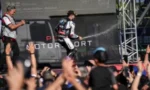 Two race drivers in racing suits on a stage, raising their hands as a cheering crowd watches; backdrop reads MOTORSPORT.