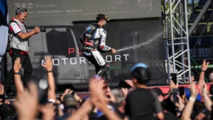 Two race drivers in racing suits on a stage, raising their hands as a cheering crowd watches; backdrop reads MOTORSPORT.