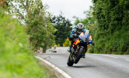 Motorcycle racer wearing teal gear leans into a sharp turn on a tree-lined country road.