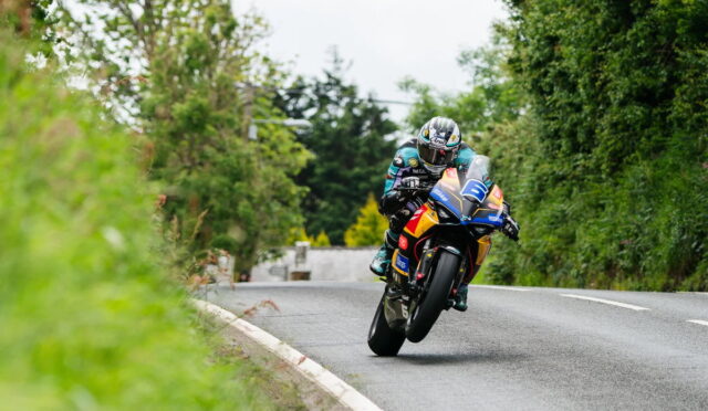 Motorcycle racer wearing teal gear leans into a sharp turn on a tree-lined country road.