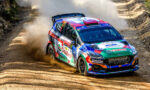 Colorful rally car corners a dusty dirt road, kicking up a cloud of dust on a rocky, forested track.