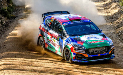 Colorful rally car corners a dusty dirt road, kicking up a cloud of dust on a rocky, forested track.