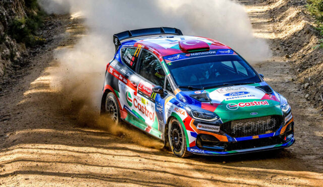 Colorful rally car corners a dusty dirt road, kicking up a cloud of dust on a rocky, forested track.
