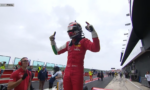 Race car driver in a red racing suit and helmet raises both arms in triumph on a pit lane with crew members nearby,