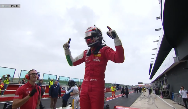 Race car driver in a red racing suit and helmet raises both arms in triumph on a pit lane with crew members nearby,