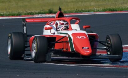 Red and white open-wheel race car with the number 10 navigates a track corner, blue and red curbs visible in the foreground.