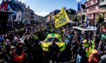 Large crowd lines a street as a bright yellow race car passes by during a rally in a European town.