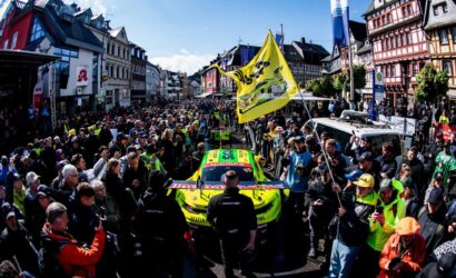 Large crowd lines a street as a bright yellow race car passes by during a rally in a European town.