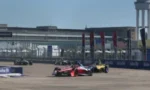 Red and black Formula-style race car leads a yellow car on a street circuit, with grandstands and a tall control tower in the background.