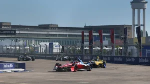 Red and black Formula-style race car leads a yellow car on a street circuit, with grandstands and a tall control tower in the background.