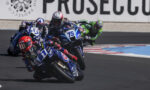 Motorcycle racers lean into a turn on a race track; blue Yamaha bikes lead the pack, with rider #75 at the front wearing a white and red helmet.