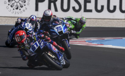 Motorcycle racers lean into a turn on a race track; blue Yamaha bikes lead the pack, with rider #75 at the front wearing a white and red helmet.