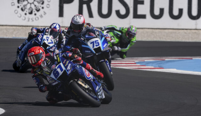 Motorcycle racers lean into a turn on a race track; blue Yamaha bikes lead the pack, with rider #75 at the front wearing a white and red helmet.