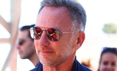 Middle-aged man wearing aviator sunglasses outdoors, close-up portrait with a reflective lens showing a red-clad figure in the reflection