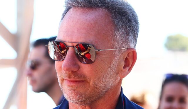 Middle-aged man wearing aviator sunglasses outdoors, close-up portrait with a reflective lens showing a red-clad figure in the reflection