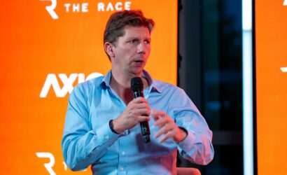 Man in a light blue shirt speaks into a handheld microphone on an orange backdrop at a panel or interview event.