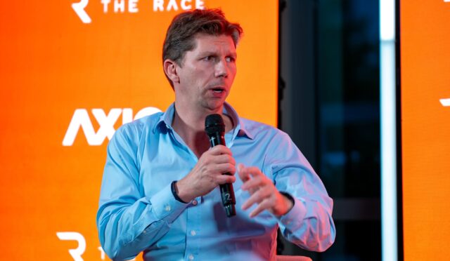 Man in a light blue shirt speaks into a handheld microphone on an orange backdrop at a panel or interview event.
