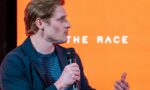 Profile view of a man in a dark blue jacket speaking into a handheld microphone on an orange stage backdrop (part of text reads 'THE RACE').