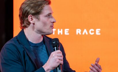 Profile view of a man in a dark blue jacket speaking into a handheld microphone on an orange stage backdrop (part of text reads 'THE RACE').
