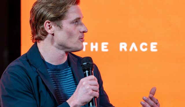 Profile view of a man in a dark blue jacket speaking into a handheld microphone on an orange stage backdrop (part of text reads 'THE RACE').