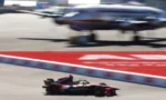 Single-seat formula race car speeds along a paved track with red markings, blurred aircraft in the background suggesting an airfield setting.