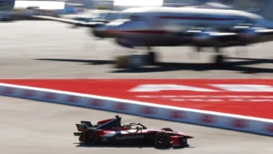 Single-seat formula race car speeds along a paved track with red markings, blurred aircraft in the background suggesting an airfield setting.