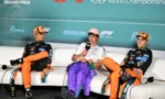 Three race car drivers in orange and purple suits sit on a white couch, mid-interview on a paddock stage with a teal banner behind them.