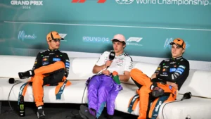 Three race car drivers in orange and purple suits sit on a white couch, mid-interview on a paddock stage with a teal banner behind them.