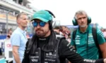 Race car driver wearing a teal cap and sunglasses in the pit lane, headset on, with team members in the background.