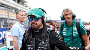 Race car driver wearing a teal cap and sunglasses in the pit lane, headset on, with team members in the background.