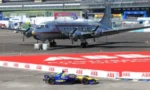 Large gray military transport aircraft on a tarmac with a blue and yellow race car in the foreground at an airshow.