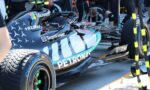 Formula 1 race car in a pit lane with sponsor logos (Microsoft, TeamViewer, Petronas) and crew members in the background inspecting the car's cockpit sideview.