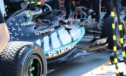 Formula 1 race car in a pit lane with sponsor logos (Microsoft, TeamViewer, Petronas) and crew members in the background inspecting the car's cockpit sideview.