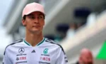 Portrait of a professional race car driver wearing a white Mercedes team polo with sponsor logos and a pink cap, standing on a racetrack with blurred grandstands in the background.