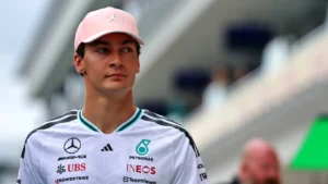 Portrait of a professional race car driver wearing a white Mercedes team polo with sponsor logos and a pink cap, standing on a racetrack with blurred grandstands in the background.