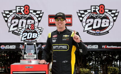 Race car driver in a SPARE Motorsports suit poses with the winner’s trophy at the FR8 Racing 208 event, Atlanta Motor Speedway backdrop.
