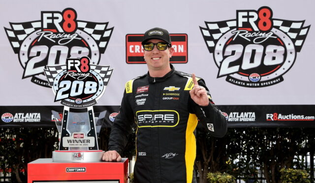 Race car driver in a SPARE Motorsports suit poses with the winner’s trophy at the FR8 Racing 208 event, Atlanta Motor Speedway backdrop.
