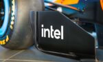 Close-up of a race car's sidepod featuring the Intel logo and bold blue‑orange bodywork, with a tire in the background, conveying branding on a Formula 1 car.
