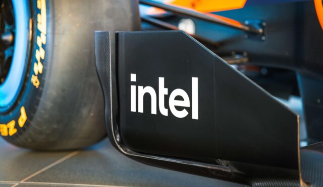 Close-up of a race car's sidepod featuring the Intel logo and bold blue‑orange bodywork, with a tire in the background, conveying branding on a Formula 1 car.