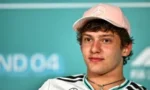 Portrait of a young man wearing a pink cap with a Mercedes logo and a white shirt with black stripes, standing in front of a teal backdrop.