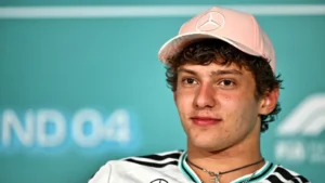 Portrait of a young man wearing a pink cap with a Mercedes logo and a white shirt with black stripes, standing in front of a teal backdrop.