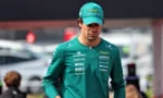 Male race car driver in a teal team suit and cap standing in a paddock area at an event.