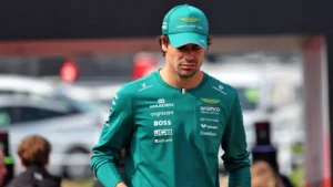 Male race car driver in a teal team suit and cap standing in a paddock area at an event.