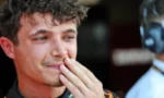 Close-up of a young man with curly dark hair touching his lips with his fingers, wearing a racing suit in a pit area.